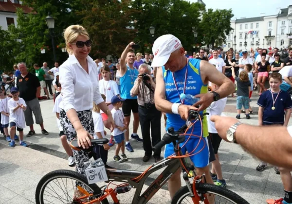 Uczestnik XVI Biegu Ulicznego NSZZ Solidarność im. Krzysztofa Zywera stoi z rowerem na placu, otoczony liczną publicznością, medalistami i fotoreporterami.