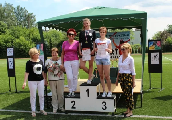 Trzy osoby stoją na podiumie podczas VI Powiatowej Olimpiady Integracyjnej w Gąbinie; dwie młode zawodniczki z medalem i dyplomem na 1. i 3. miejscu, otoczone przez organizatorów pod zielonym namiotem na tle boiska.