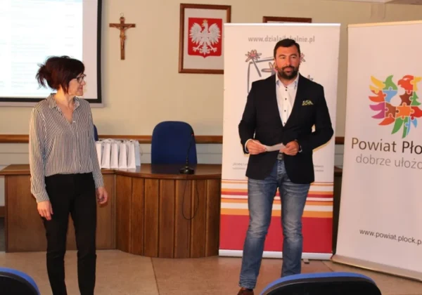 Mężczyzna w czarnej marynarce i jeansach przemawia podczas prezentacji, stojąc obok kobiety w paskowanej koszuli; w tle widoczne banery Powiatu Płockiego i krzyż w sali konferencyjnej.