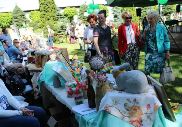 Grupa osób ogląda stoły z rękodziełem, kwiatami i dekoracjami podczas Dnia Integracji i Aktywności Osób Niepełnosprawnych na świeżym powietrzu.