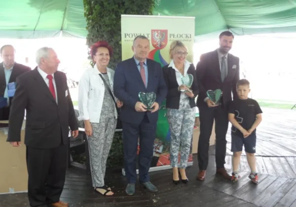 Grupa uczestników stoi na scenie pod zielonym namiotem podczas jubileuszowego XV Festynu z Nadzieją, trzymając kryształowe serca jako trofea, z bannerem Powiatu Płockiego w tle.