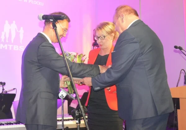 Trzy osoby w garniturach stoją na scenie podczas jubileuszu XX-lecia MTBS Sp. z o.o., gdy mężczyzna wręcza kwiaty kobiecie.