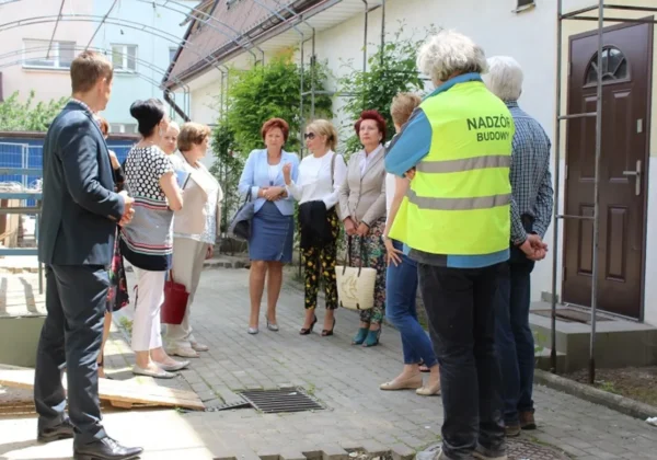 Grupa urzędników i gości stoi na placyku przed wejściem do budynku podczas wyjazdowego posiedzenia Komisji do Spraw Społecznych; wśród nich osoba w żółtej kamizelce odblaskowej z napisem NADZÓR BUDOWY.