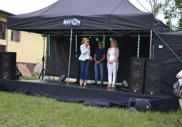 Trzy osoby stoją na scenie pod czarnym namiotem podczas festynu rodzinnego w Bielsku; jedna kobieta przemawia do mikrofonu, a dwie pozostałe stoją obok, z głośnikami i oświetleniem w tle.