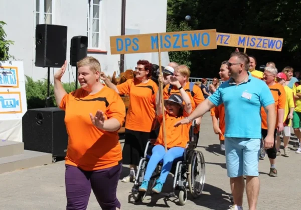 Uczestnicy XXIV Letniej Olimpiady Sportowej Nowe Miszewo 2016 maszerują z banerem DPS MISZEWO, wielu w pomarańczowych koszulkach, w tym osoba na wózku inwaliskim.
