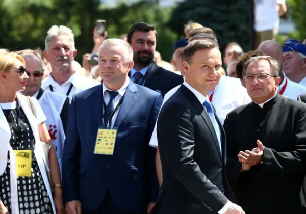 Grupa osób w garniturach oraz duchowny w sutannie stoi na zewnątrz podczas uroczystości, wszyscy z żółtymi identyfikatorami zawieszonymi na szyjach.