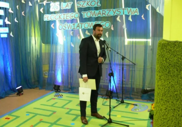 Mężczyzna w garniturze stoi przy mikrofonie na szkolnej scenie, a w tle wisi niebieska zasłona i baner z napisem „25 lat szkół Płockiego Towarzystwa Oświatowego”.