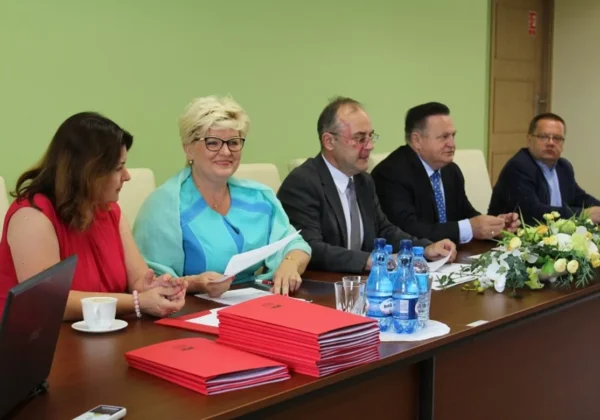 Grupa osób w formalnych strojach siedzi przy stole konferencyjnym podczas posiedzenia Konwentu Państwowej Wyższej Szkoły Zawodowej w Płocku, z czerwonymi teczkami, butelkami wody i bukietem kwiatów na stole.