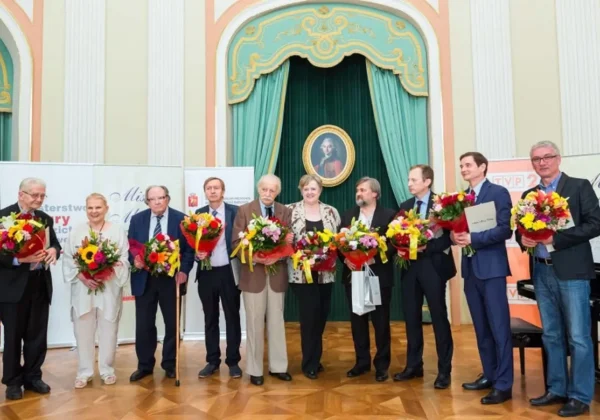 Grupa laureatów XVI edycji Programu Mistrz Mowy Polskiej stoi na scenie i trzyma bukiety kwiatów, na tle ozdobnej sali z zielonymi zasłonami i portretem w ramie.