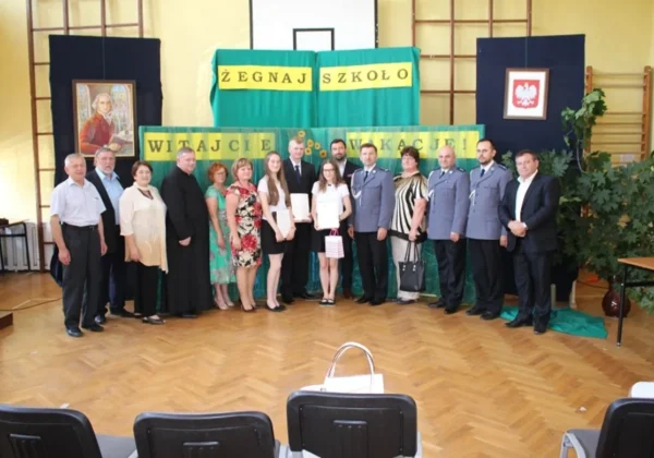 Grupa osób stojących w szkolnej sali gimnastycznej przed dekoracją z napisami "Żegnaj szkoło" i "Witajcie wakacje", wśród nich uczniowie z nagrodami i dorosli w strojach galowych.