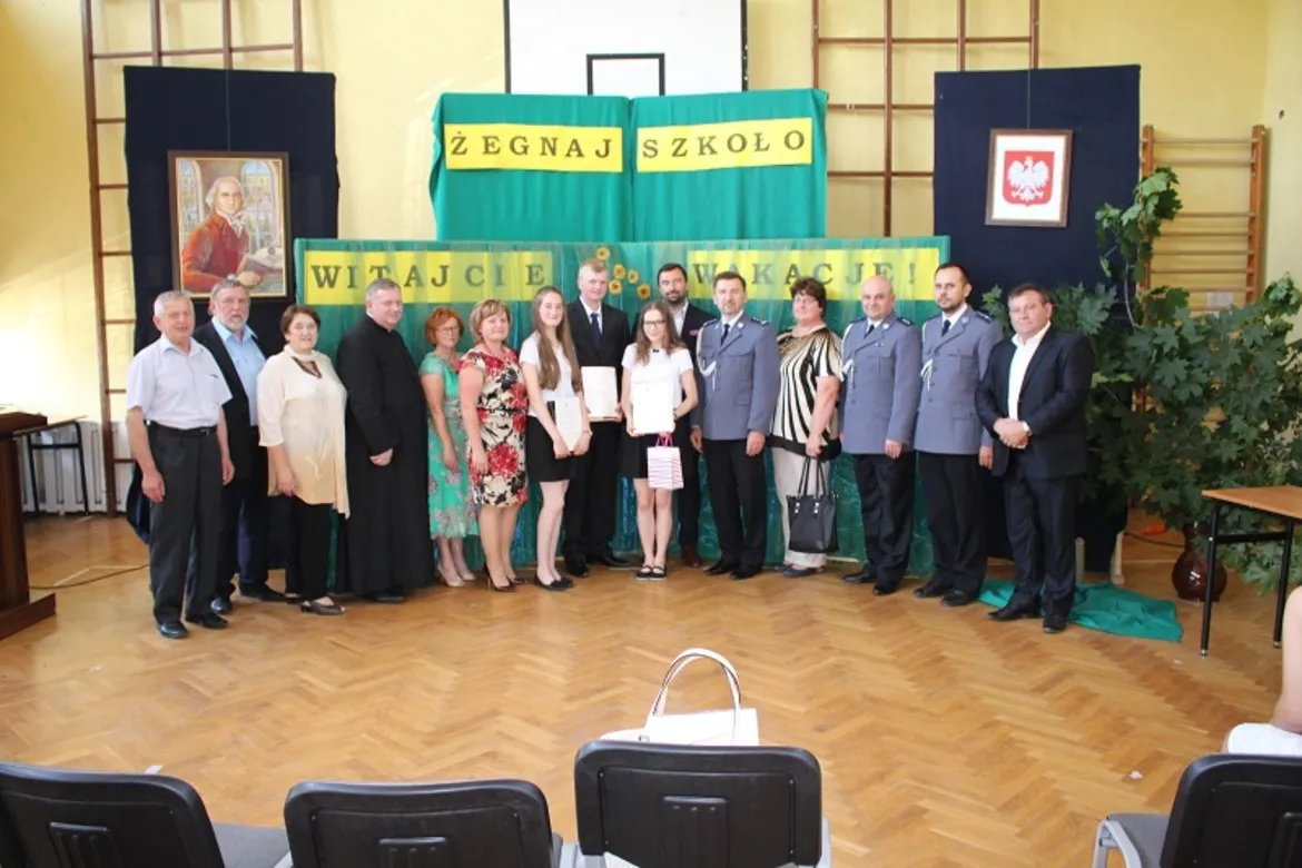 Grupa osób stojących w szkolnej sali gimnastycznej przed dekoracją z napisami "Żegnaj szkoło" i "Witajcie wakacje", wśród nich uczniowie z nagrodami i dorosli w strojach galowych.