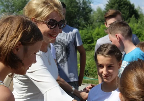 Dorosła kobieta w białej bluzce rozmawia z młodą dziewczynką podczas pikniku integracyjnego dla rodzin zastępczych i prowadzących Rodzinne Domy Dziecka na tle zieleni.