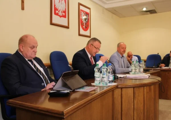 Grupa radnych podczas sesji Rady Powiatu siedzi przy drewnianym, zakrzywionym stole z mikrofonami, dokumentami i butelkami wody, a na ścianie widoczne są herby.