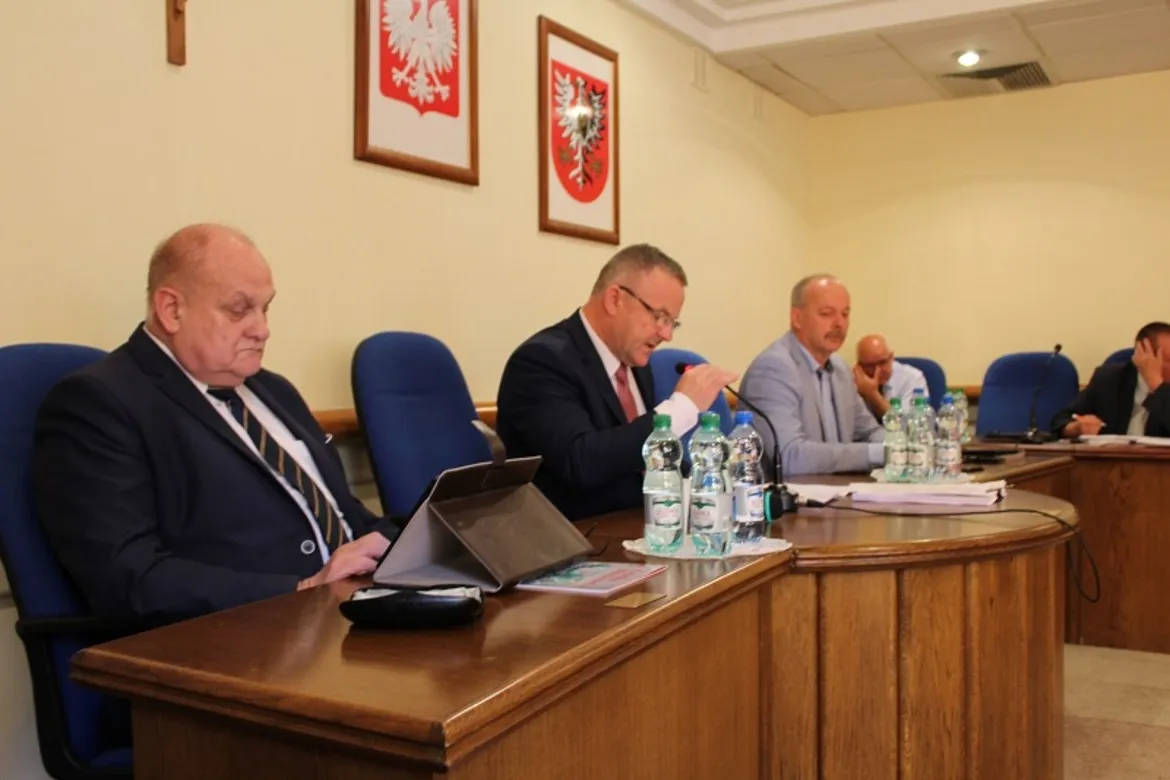 Grupa radnych podczas sesji Rady Powiatu siedzi przy drewnianym, zakrzywionym stole z mikrofonami, dokumentami i butelkami wody, a na ścianie widoczne są herby.