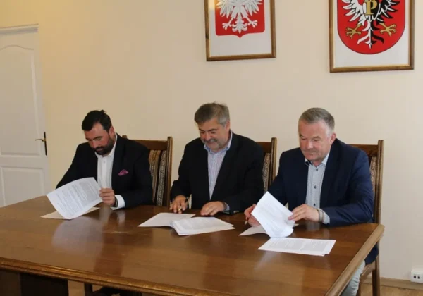Trzech mężczyzn w garniturach podpisuje dokumenty przy stole podczas podpisywania kolejnej umowy na remonty dróg powiatu płockiego.
