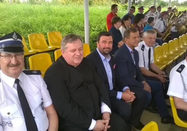 Grupa mężczyzn w mundurach strażackich i garniturach siedzi na żółtych krzesełkach podczas Miejsko-Gminnych Zawodów Sportowo-Pożarniczych, z orkiestrą w tle.