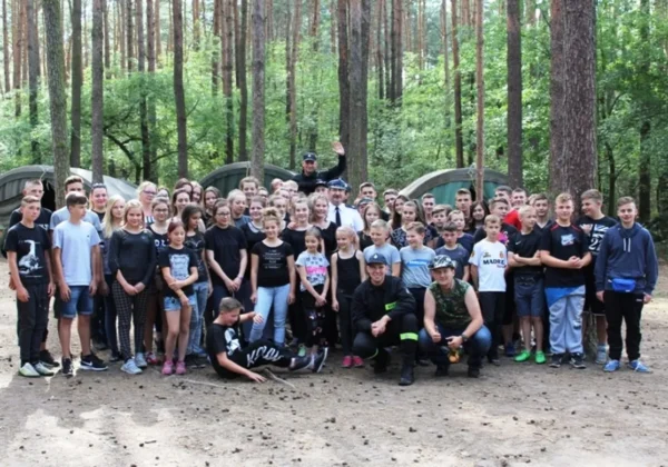 Duża grupa młodych strażaków z powiatu płockiego na obozie Młodzieżowych Drużyn Pożarniczych, stojąca razem przed namiotami w lesie.