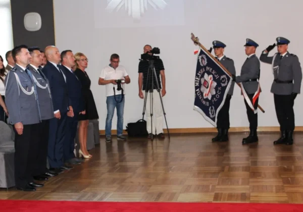 Uroczystość Święta Policji: grupa funkcjonariuszy i urzędników stoi w sali, po prawej trzech policjantów w mundurach trzyma sztandar i salutuje, w tle kamerzyści i fotoreporterzy.
