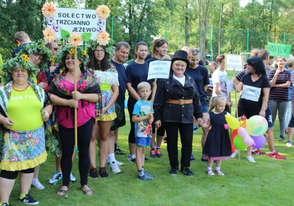 Grupa mieszkańców podczas VI Gminnego Turnieju Sołectw w Nowym Duninowie stoi na zielonej trawie, trzymając transparenty z nazwami sołectw i balony.