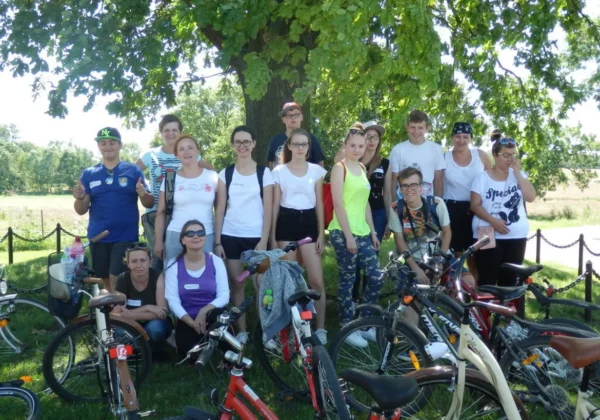 Grupa młodych ludzi i dorosłych stoi pod dużym drzewem, otoczona rowerami — początek rajdu rowerowego promującego historię Gminy Bulkowo łączącej pokolenia.