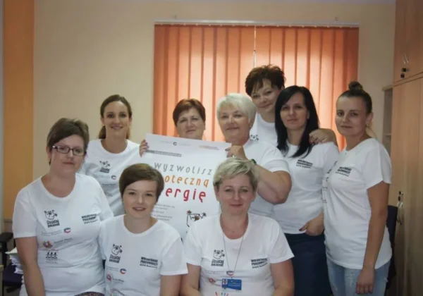 Grupa wolontariuszek w białych koszulkach z logo stoi w sali w gminie Bodzanów, trzyma plakat i uśmiecha się do zdjęcia.