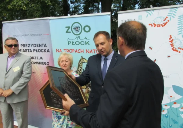Kilka dorosłych osób w garniturach stoi na tle bannerów ZOO Płock podczas wręczania pamiątkowej ramy z okazji święta pszczelarzy i gratulacji od Powiatu Płockiego.