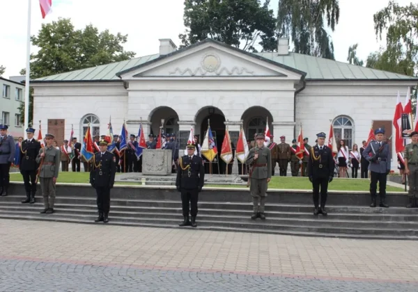 Grupa uczestników w mundurach i strojach cywilnych stoi na schodach przed zabytkowym budynkiem, trzymając flagi podczas patriotycznej ceremonii upamiętniającej Bohaterów 1920 roku w Płocku.