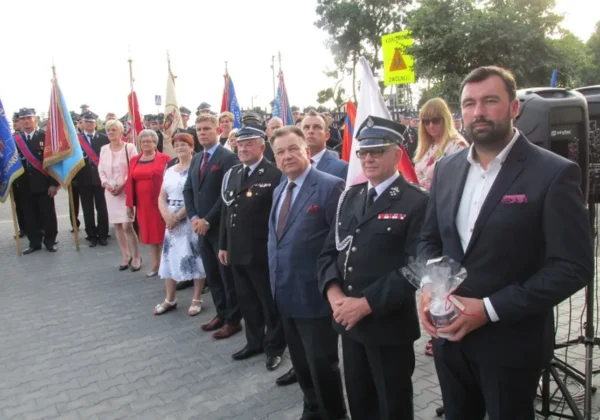 Grupa osób w mundurach strażackich i eleganckich garniturach stoi na placu podczas obchodów 60-lecia OSP w Słupnie, z flagami w tle.