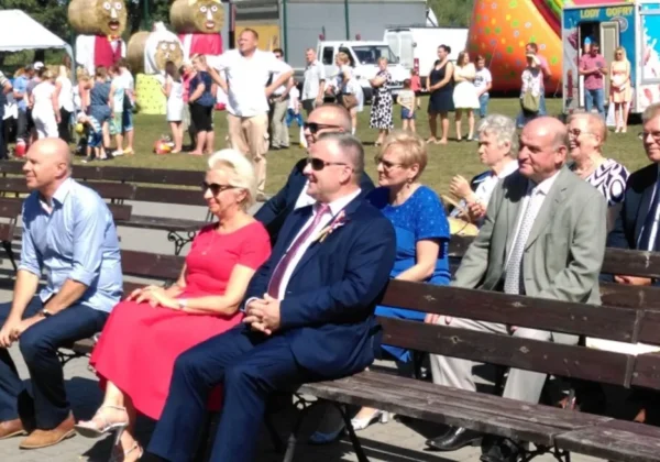 Grupa dorosłych siedzi na ławkach podczas Święta plonów w gminach Stara Biała i Brudzeń Duży, z tłumem ludzi i atrakcjami w tle.