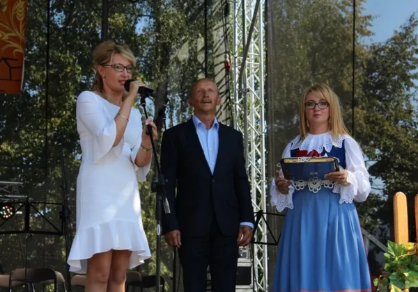 Trzy osoby stoją na scenie podczas Dożynek Gminnych w Bielsku: wokalistka w białej sukni śpiewa do mikrofonu, mężczyzna w garniturze stoi między nią a kobietą w ludowym stroju trzymającą ozdobne niebieskie pudełko.