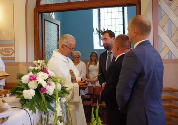 Ksiądz w białej sutannie wręcza przedmiot grupie mężczyzn w garniturach podczas Dożynek Parafialnych w Radzanowie i Święta Plonów w Blichowie, w kościelnym wnętrzu przy dekoracyjnym bukiecie kwiatów.