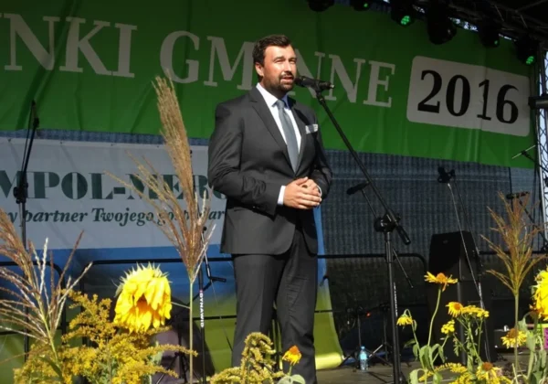 Mężczyzna w garniturze przemawia ze sceny podczas Święta Plonów Gminy Drobin, na tle zielonego baneru z napisem 2016 i dekoracji ze zbóż i kwiatów.