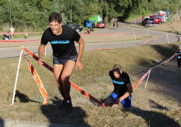 Dwóch uczestników aquathlonu w czarnych koszulkach z logo AQUATHLON wspina się pod stromą skarpą na trasie w Płocku, otoczonych pomarańczowymi taśmami i widzami w tle.