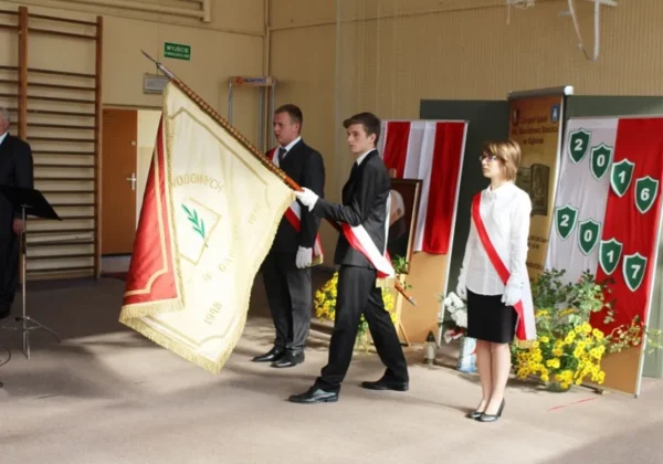 Uczniowie w garniturach i białych koszulach niosą sztandar szkoły podczas uroczystego apelu, na tle dekoracji w barwach narodowych i kwiatów.