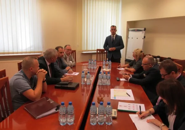 Grupa osób w sali konferencyjnej siedzi przy długim stole, a stojący w garniturze mężczyzna prowadzi spotkanie przy flip chart, wokół leżą notatniki, dokumenty i liczne butelki wody.