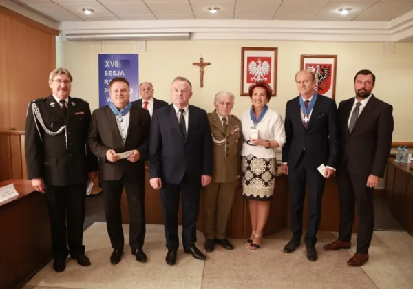 Ośmiu uczestników w garniturach i mundurze z medalami na szyi stoi w sali sesyjnej, w tle plakat XVII Sesja oraz polskie godła.