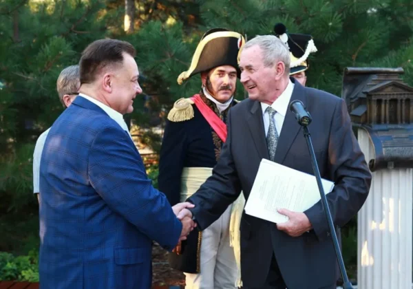 Dwóch mężczyzn w garniturach ściska sobie dłonie podczas uroczystego odsłonięcia pomnika upamiętniającego powstanie Teatru w Płocku w 1912 roku, w tle widoczne osoby w historycznych mundurach i tablica pamiątkowa.