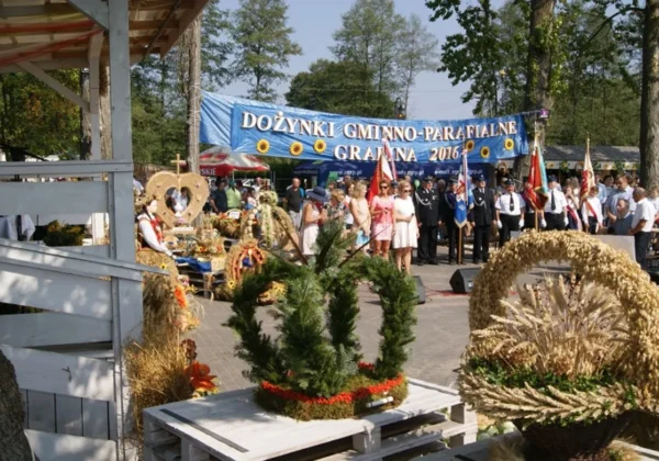 Uroczystość Dożynki Gminno-Parafialne w gminie Łąck, na placu dekorowane wieńce żniwne i grupa osób w strojach ceremonialnych pod dużym banerem z napisem Dożynki Gminno-Parafialne.