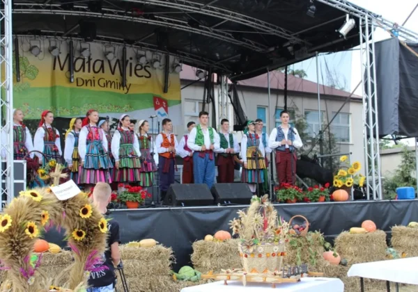 Grupa wykonawców w kolorowych strojach ludowych występuje na scenie podczas Dni Małej Wsi i Dożynek Gminnych, a przed sceną widoczne są jesienne dekoracje z kwiatów, siana i dyni.