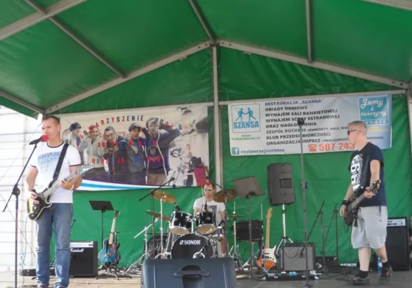 Na scenie pod zielonym namiotem trójka muzyków gra na gitarze, perkusji i basie, a w tle widoczne są banery z hasłem „Nie Ćpam – Nie Piję – Teraz Żyję!”.