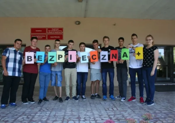 Grupa młodzieży stojąca przed wejściem do szkoły trzyma kolorowe kartoniki z literami tworzące hasło BEZPIECZNA+.