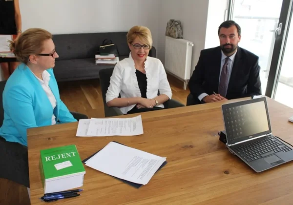 Trzy osoby siedzą przy stole w biurze, omawiają plany Powiatu Płockiego dotyczące Hospicjum i Zakładu Opiekuńczo-Leczniczego; na stole leżą dokumenty, teczki i laptop.