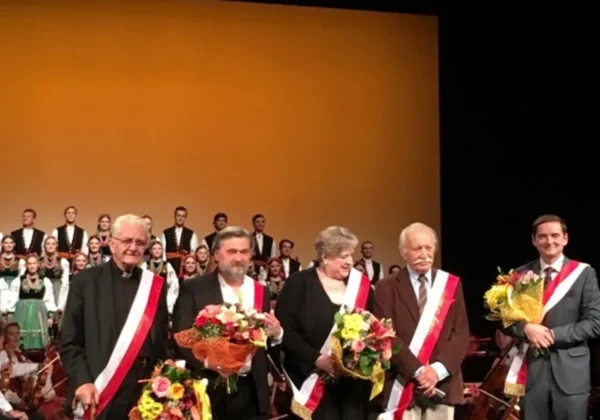 Na scenie pięć osób w eleganckich strojach z biało-czerwonymi wstęgami trzyma bukiety kwiatów, a w tle stoi chór na żółto-pomarańczowym tle.