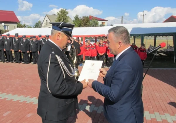 Uroczyste wręczanie dyplomu i pucharu podczas jubileuszu 50-lecia OSP Rogozin, na tle strażaków w mundurach i czerwonych kurtkach pod plenerowym namiotem.