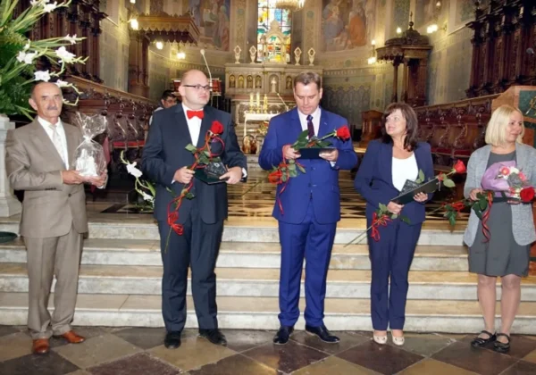 Pięć osób w formalnych ubraniach stoi na schodach kościoła, trzymając statuetki i kwiaty podczas wręczania podziękowań.