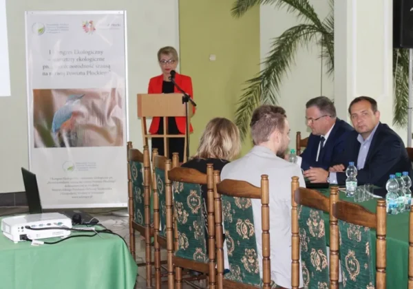 Prelegentka w czerwonym żakiecie wygłasza przemówienie z mównicy podczas kongresu o bioróżnorodności w Powiecie Płockim, na tle plakatu i zgromadzonych uczestników.