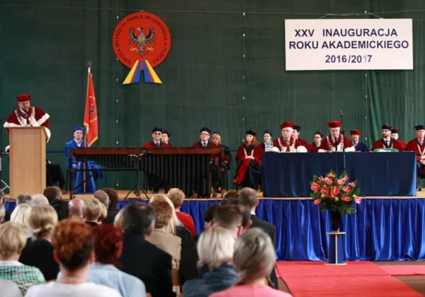 Przewodniczący Rady Powiatu przemawia z podium podczas XXV inauguracji roku akademickiego 2016/2017, na scenie z członkami senatu w togach i gośćmi, przed zgromadzoną publicznością.