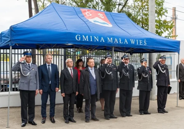 Grupa strażaków i urzędników w garniturach stoi pod niebieskim namiotem z herbem i napisem „Gmina Mała Wieś”, salutując podczas uroczystości z okazji rozszerzenia Krajowego Systemu Ratowniczo-Gaśniczego.