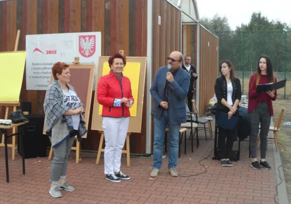 Grupa osób na zewnątrz podczas Pikniku Integracyjnego „Bezpieczna +”, mężczyzna z mikrofonem przemawia, obok stoją kobiety, w tle tablice edukacyjne na stojakach.