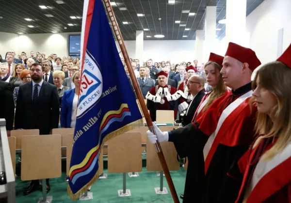 Uroczystość inauguracji roku w PWSZ, na pierwszym planie grupa studentów w czerwonych togach niesie sztandar, w tle zgromadzeni goście.